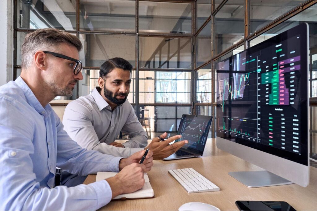 Two businessmen analyzing stock market data on computer screens in an office setting, discussing financial trends and making decisions.